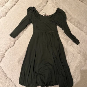 Mörkgrön klänning från Miss Sixty - Supersnygg mörkgrön klänning från Miss Sixty med trekvartsärm och puffiga axlar. Klänningen har en markerad midja och ett mjukt, följsamt tyg som ger ett fint fall. Perfekt för dig som gillar en feminin och trendig look.
