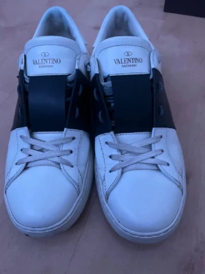 Valentino Garavani vita sneakers - Snygga vita sneakers från Valentino Garavani med bred svart läderdetalj över snörningen och klassisk logga på plösen. Skorna har lågt skaft och är tillverkade i slätt läder med vita snören och diskreta nitar på hälen. Perfekta för en clean och stilren look.
