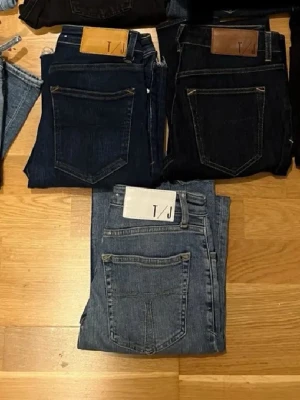 Blå och svarta jeans från T/J - Säljer flera par jeans från T/J i olika blå och svarta nyanser. Klassisk femficksmodell med raka ben och tydliga sömmar. Jeansen har läderpatch med logga bak i midjan och är tillverkade i denim. Perfekta för en stilren och avslappnad look.