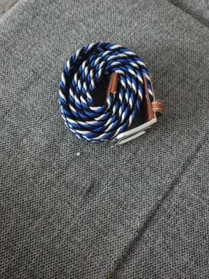 Flätat bälte Grisch - Snyggt flätat bälte med randigt mönster i blått, vitt och svart. Bältet har detaljer i brunt skinn och ett silvrigt spänne. Perfekt accessoar för att lyfta din outfit.