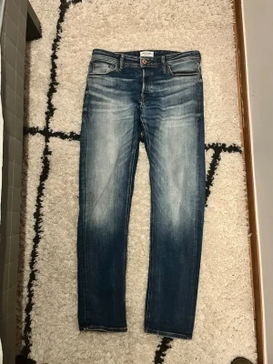 Blå slim fit jeans från Jack and jones - Säljer ett par blå slim fit jeans från Jack and Jones med snygga slitningar och ljusa partier framtill och bak. Klassisk femficksmodell med dragkedja och knapp. Perfekta för en avslappnad och trendig look.