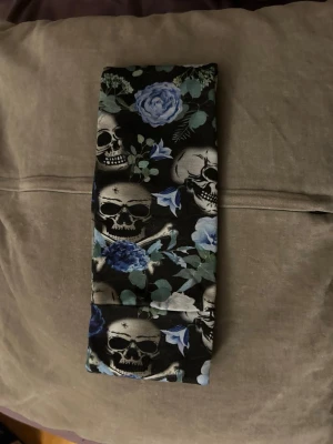 Svart hårband med dödskallar och blommor - Snyggt svart hårband med tryck av vita dödskallar och blå rosor samt gröna blad. Perfekt accessoar för dig som gillar alternativ stil och vill sticka ut. Mjukt och stretchigt material som sitter bekvämt i håret.