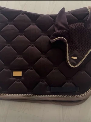 Brunt schabrak och huva med kantband - Säljer ett mörkbrunt schabrak med matchande huva till häst. Båda har quiltat mönster och dekorativt kantband i beige och guld. Schabraket har rektangulär form och detaljer i guld. Materialet är vadderat tyg och huvan är virkad med tyg vid öronen.