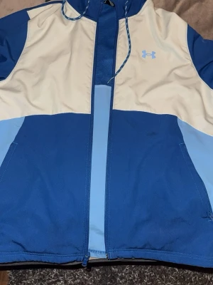 Blå och vit vindjacka från Under Armour - Snygg vindjacka från Under Armour i blått och vitt med dragkedja framtill och huva med snörning. Jackan har två sidofickor och är perfekt för dig som gillar färgglada kläder. Loggan syns på bröstet och materialet känns lätt och funktionellt. Jackan är i nytt skick bara använd 3-4 gånger. 
