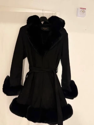 Svart kappa med fuskpälsdetaljer - Stilren svart kappa från Gorgeous med bred krage, ärmslut och nederkant i mjuk fuskpäls. Kappan har knytband i midjan för en snygg siluett och är helsvart för en klassisk look. Perfekt för dig som vill ha en trendig och varm ytterjacka.