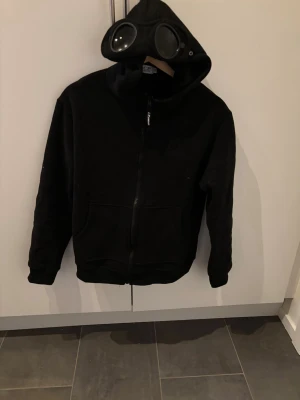C.P company hoodie - Säljer en svart hoodie från C.P. Company med dragkedja och ikoniska goggles i huvan. Hoodien har två fickor framtill och broderad logga på bröstet. Tillverkad i mjukt material som känns skönt mot huden. Perfekt för dig som vill sticka ut med en unik streetwear-look.