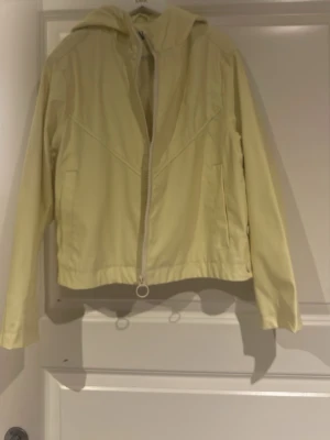 Ljusgul vindjacka/regnjackq med huva - Snygg ljusgul vindjacka med dragkedja och huva. Jackan har raka ärmar, två sidofickor och en enkel, stilren design. Perfekt för blåsiga dagar och lätt att matcha med olika outfits. Materialet känns tunt och smidigt. Storlek 152
