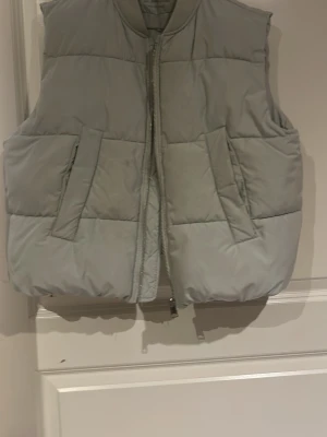 Ljusgrå quiltad dunväst - Stilren ljusgrå dunväst med quiltad design och två stora snedställda fickor framtill. Västen har hög krage och dragkedja hela vägen. Har dock en liten fläck som går att tvära bort. Storlek 164