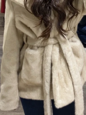 Beige fuskpälsjacka med knytskärp - Snygg beige fuskpälsjacka med bred krage och knytskärp i midjan. Jackan har en mjuk och fluffig känsla, långa ärmar och två fickor framtill. Perfekt för dig som vill ha en trendig och varm look under kalla dagar.