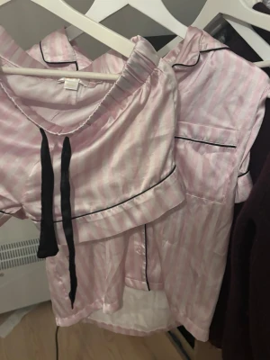 Rosa randig pyjamas med svarta detaljer - Söt pyjamas i ljusrosa och vita ränder med svarta kontrastdetaljer. Setet består av kortärmad skjorta med krage och bröstficka samt matchande shorts med svart dragsko. Vs dupe