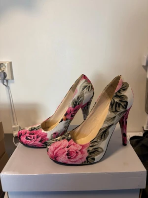 Blommiga pumps med hög klack - Säljer ett par klackar med blommigt mönster i rosa, grönt och vitt. Skorna har rundad tå och hög ungefär 8-10 cm klack. Materialet är textil med tryckt blommönster. 