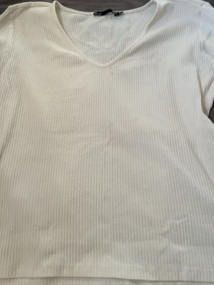 Vit ribbad v-ringad topp - Vit ribbad topp med v-ringning och korta ärmar. Tunn och stretchig i materialet, perfekt för en enkel och clean look. Passar till det mesta och är lätt att styla.