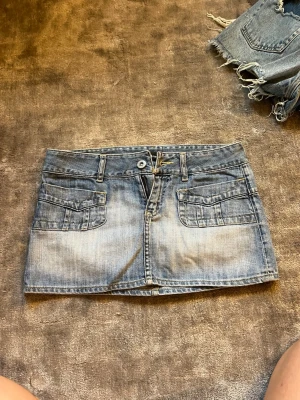 Blå jeanskjol från Dolce Ribelle - Säljer en kort blå jeanskjol från Dolce Ribelle med två stora fickor fram och bälteshällor. Kjolen har klassisk denimlook och stängs med knapp och dragkedja. Perfekt för dig som gillar en avslappnad och trendig stil. Lågmidjad. Motsvarar storlek 36