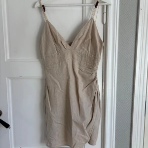 Beige kort klänning med v-ringning - Säljer en beige kort klänning med v-ringning och smala axelband. Klänningen har en figurnära passform och är tillverkad i ett linneliknande material. Perfekt för varma dagar och enkel att styla med accessoarer.