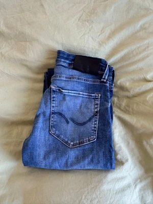 Blå slim glen jeans, Jack & Jones 28:30 - Snygga blå slim fit jeans från Jack & Jones med klassisk femficksdesign och tvättande design. Jeansen har en mörk tvätt och svarta detaljer vid linningen. Perfekta för en avslappnad och trendig stil.