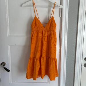 Orange kort klänning med volang - Snygg orange kort klänning med tunna axelband och v-ringning. Klänningen har broderade detaljer och volang nedtill för en lekfull look. Perfekt för varma dagar och sommarens alla tillfällen.