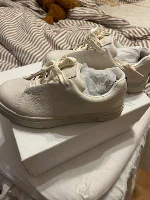 Vita sneakers från Eytys i läder - Stilrena vita sneakers från Eytys med klassisk låg profil och rund tå. Skorna är tillverkade i mjukt, strukturerat läder och har matchande vita skosnören. Endast använda en gång, säljer då de är lite för små!