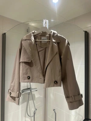 Kort beige trenchcoat från Bershka - Trendig kort trenchcoat i beige från Bershka. Jackan har bred krage, dubbelknäppning med stora knappar och justerbara spännen vid ärmsluten. Tillverkad i ett lätt bomullsliknande material, perfekt för lager på lager och en cool streetstyle-look.