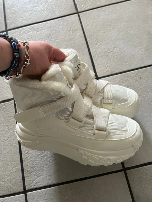 Vita quiltade moon boots med fuskpäls storlek 40 - Säljer ett par vita moon boots med quiltad ovandel, breda kardborreband och fluffigt fuskpälsfoder. Skorna har en chunky sula och hög design som håller värmen. Perfekta för kalla vinterdagar och riktigt snygga till streetwear-looken.