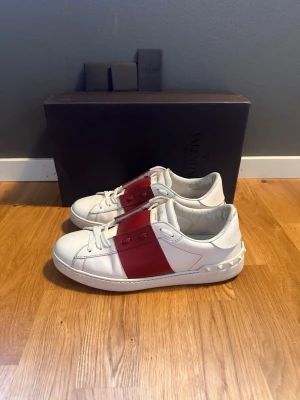 Valentino vita sneakers med röd detalj - Snygga vita sneakers från Valentino med bred röd läderdetalj över mitten och ikoniska nitar på hälen. Skorna har klassisk låg profil, vita snören och en chunky sula. Perfekta för dig som vill ha en stilren men ändå unik look. Size 41, bra skick 