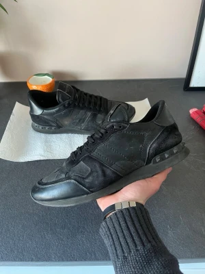 Svarta Valentino Garavani sneakers - skick 7/10. Säljer ett par svarta Valentino Garavani sneakers med coola nitar på hälen. Skorna har en mix av mocka och läderdetaljer, samt svarta skosnören och en chunky sula. Perfekta för dig som gillar exklusiva och stilrena sneakers.