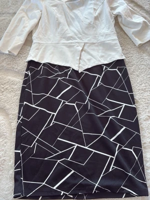 Svartvit långklänning med grafiskt mönster - Snygg långklänning med vit överdel och svart kjol med vitt grafiskt mönster. Klänningen har trekvartsärm, markerad midja och dragkedja i ryggen. Perfekt för dig som gillar stilrena kontraster och modern design.