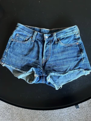 Levi's blå jeansshorts hög midja - Klassiska blå jeansshorts från Levi's med en ganska lågmidja och råa kanter. Femficksmodell med knapp och dragkedja framtill samt Levi's ikoniska läderpatch bak. Perfekta för sommaren och har en avslappnad, loose passform.