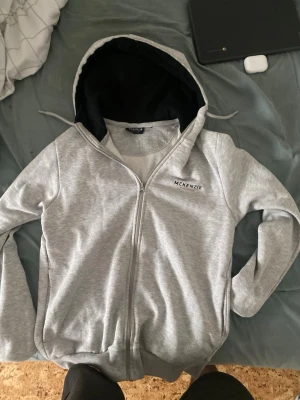 Grå zip hoodie från McKenzie - Snygg grå zip hoodie från McKenzie med svart innerfoder i huvan och diskret logga på bröstet. Hoodien har dragsko i huvan, långa ärmar och ribbade muddar. Perfekt för en chill och avslappnad stil. Materialet känns mjukt och bekvämt, troligen bomullsblandning.