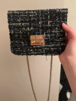 Svart tweed kuvertväska med kedja - Snygg kuvertväska i svart tweed med inslag av blått och silver, dekorerad med små paljetter. Väskan har guldfärgad kedjerem och ett guldfärgat lås framtill. Invändigt finns ett fack med dragkedja. Perfekt accessoar för att lyfta din outfit. Pris kan diskuteras 