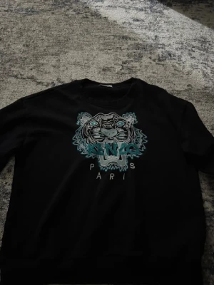 Svart sweatshirt med tiger från Kenzo - Svart sweatshirt från Kenzo med ikonisk broderad tiger i blått, vitt och turkost på bröstet samt texten 'KENZO PARIS'. Tröjan har rund halsringning och är tillverkad i mjukt bomullsmaterial. Perfekt för dig som gillar streetwear och vill sticka ut.