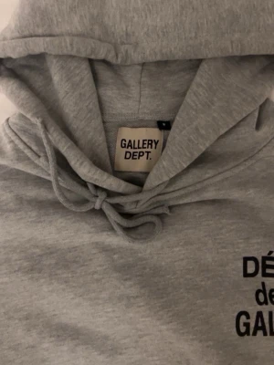 Grå hoodie från Gallery Dept. - Säljer en ljusgrå hoodie från Gallery Dept. med svart tryck på bröstet. Hoodien har huva med dragsko och är tillverkad i mjukt sweatshirtmaterial. Perfekt för en iavslappnad och trendig stil.