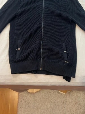 Mörkblå ziptröja från Calvin Klein - Snygg mörkblå stickad ziptröja från Calvin Klein med ribbad krage och två fickor framtill med knappdetaljer. Tröjan har en hel dragkedja och diskret logga på ena fickan. Perfekt för lager på lager och enkel att matcha.