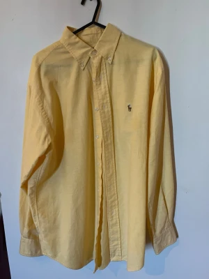 Gul skjorta från Ralph Lauren - Klassisk gul skjorta från Ralph Lauren med button-down krage och broderad logga på bröstet. Skjortan har långa ärmar, knappar framtill och är tillverkad i mjuk bomull. Perfekt för en clean och stilren look. STORLEK M