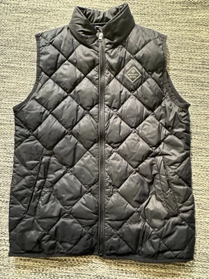 Svart quiltad dunväst från GANT - Snygg svart dunväst från GANT med quiltat mönster och hög krage. Västen har dragkedja framtill, två sidofickor och en diskret logotyp på bröstet. Perfekt för lager på lager och ger en clean look till din outfit.