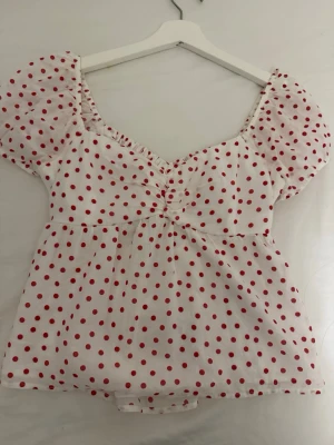 Vit prickig blus från Hollister - Supersöt vit blus från Hollister med röda prickar och puffärmar. Blusen har en lätt rynkad detalj vid bysten och en feminin v-ringning. Perfekt för dig som gillar en lekfull och trendig stil.
