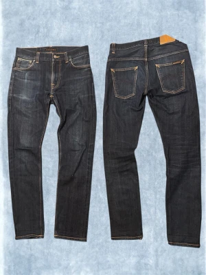 NUDIE JEANS (SELVAGE) - Snygga mörkblå jeans från Nudie med smal passform och klassiska kontrastsömmar. Jeansen har fem fickor, gylf med knapp och dragkedja samt Nudie-logga på bakfickan. Tillverkade i slitstark denim, perfekta till vardagslooken.