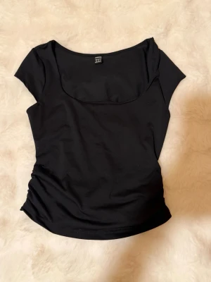 Svart topp med rund hals från SHEIN - En enkel och stilren svart topp från SHEIN med rundad halsringning och korta ärmar. Toppen är figurnära och tillverkad i ett mjukt syntetmaterial som känns bekvämt mot huden. Perfekt att matcha med jeans eller kjol.