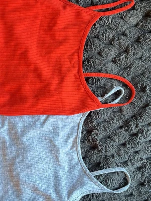 Två ribbade linnetoppar i rött och grått - Säljer två stilrena linnetoppar med smala axelband, en i klarrött och en i ljusgrått. Båda topparna är ribbade och har en enkel, croppad passform som passar perfekt till jeans eller kjol. Materialet känns mjukt och stretchigt, troligen bomull eller bomullsblandning.