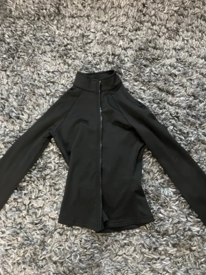 Svart sportig jacka från Dobsom - Svart, sportig jacka från Dobsom med hög krage och hel dragkedja framtill. Jackan har en slim passform och är tillverkad i ett mjukt syntetmaterial som passar perfekt för träning eller som ett extra lager. Långärmad och enkel design utan synliga mönster.