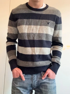 Randig stickad tröja från American Eagle - Snygg stickad tröja från American Eagle med breda horisontella ränder i mörkblått, grått och ljusgrått. Tröjan har rund halsringning, ribbade muddar och en liten broderad logga på bröstet. Perfekt för dig som gillar klassisk och avslappnad stil.
