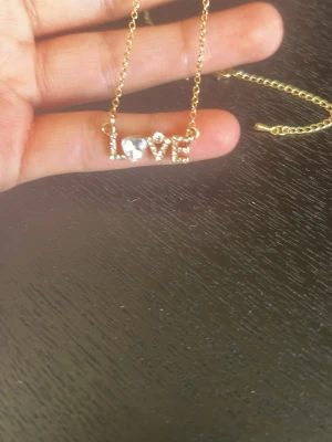 Guldigt halsband med LOVE och pärla - Snyggt halsband i guldfärgad metall med ett hänge som stavar ut 'LOVE' dekorerat med små glittrande stenar och en vit pärla som ersätter O:et. Kedjan är tunn och elegant, perfekt för att ge en söt touch till din outfit.