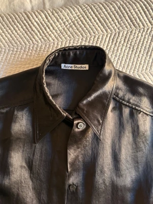 Svart silkesliknande skjorta Acne Studios - Snygg svart skjorta från Acne Studios i ett glansigt, silkesliknande material. Klassisk krage, knäppning framtill och långa ärmar. Perfekt för dig som vill ha en stilren och modern look.
