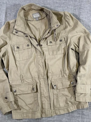 Converse One Star khaki field jacket XL military utility workwear - Converse One Star field jacket XL khaki. Military-inspired utility jacket with flap pockets, snap + zip front, quilted shoulders, adjustable cuffs, and stand collar with buckle detail. Missing waist belt but still keeps a clean silhouette. Durable 100% cotton-great for workwear or everyday layering. Good used condition, light wear + small spot on right collar (see photo).  Measurements: Waist 50, P2P 54, S2S 37, Length 67, Sleeve 61 cm