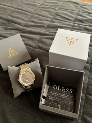 Guess klocka i silver och guld - Stilren Guess klocka med länkarmband i silver och guld. Dekorerad med gnistrande stenar och har romerska siffror. Klockan levereras i originalask med tillhörande manual. Äkthetsbevis ingår.