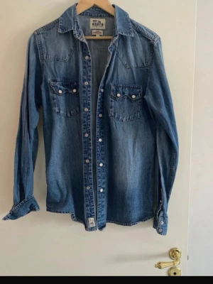Blå denimskjorta  - Säljer en klassisk blå denimskjorta lång ärm och snygga knappar framtill. Skjortan har två bröstfickor med knappstängning och en cool western-inspirerad söm på ryggen. Perfekt för dig som gillar en avslappnad och tidlös look.