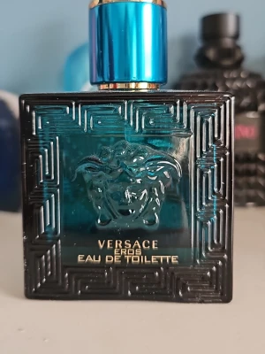 Versace Eros Eau de Toilette - Versace Eros Eau de Toilette i en stilren, fyrkantig flaska i turkost glas med svarta detaljer och guldtext. Flaskan har ett klassiskt Versace-mönster och Medusa-huvud i relief på framsidan samt en metallisk blå kork. En ikonisk doft för dig som gillar lyxiga parfymer.