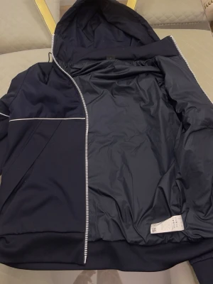 Mörkblå vindjacka med huva - Snygg mörkblå vindjacka med hel dragkedja och huva. Jackan har vita detaljer i form av tunna linjer över bröst och rygg samt längs ärmarna. Tillverkad i ett lätt och vindtätt material, perfekt för blåsiga dagar. Praktiska fickor framtill och enkel, stilren design.Den e exakt som ny har använt i 1 vecka bara. Storlek M