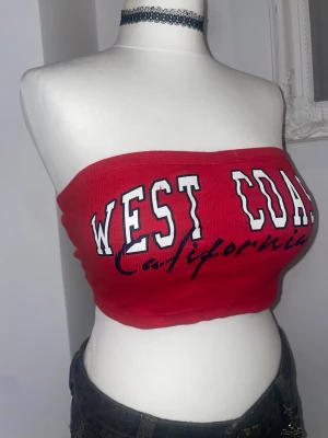 Röd bandeau tubtopp - Snygg röd bandeau tubtopp med texten 'West Coast California' i vitt och svart framtill. Toppen är axelbandslös och croppad. Storlek S men passar mer som en M. 96% bomull 