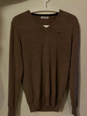 Beige V-ringad tröja J.Lindeberg - Snygg beige stickad tröja från J.Lindeberg med V-ringning och diskret logga på bröstet. Tillverkad i tunt, mjukt material som passar perfekt till lager på lager. Långa ärmar och ribbade muddar för en clean look.