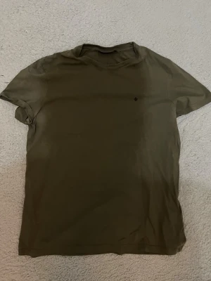 Olivgrön t-shirt med liten logga - En enkel olivgrön t-shirt med rund hals och korta ärmar. Liten diskret logga på bröstet. Perfekt basplagg som funkar till det mesta. Tillverkad i mjuk bomull för skön känsla hela dagen.